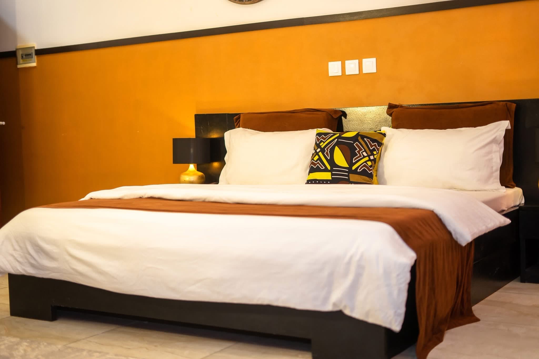 Chambre au mur ocre avec grand lit, linge blanc immaculé et coussin aux motifs africains